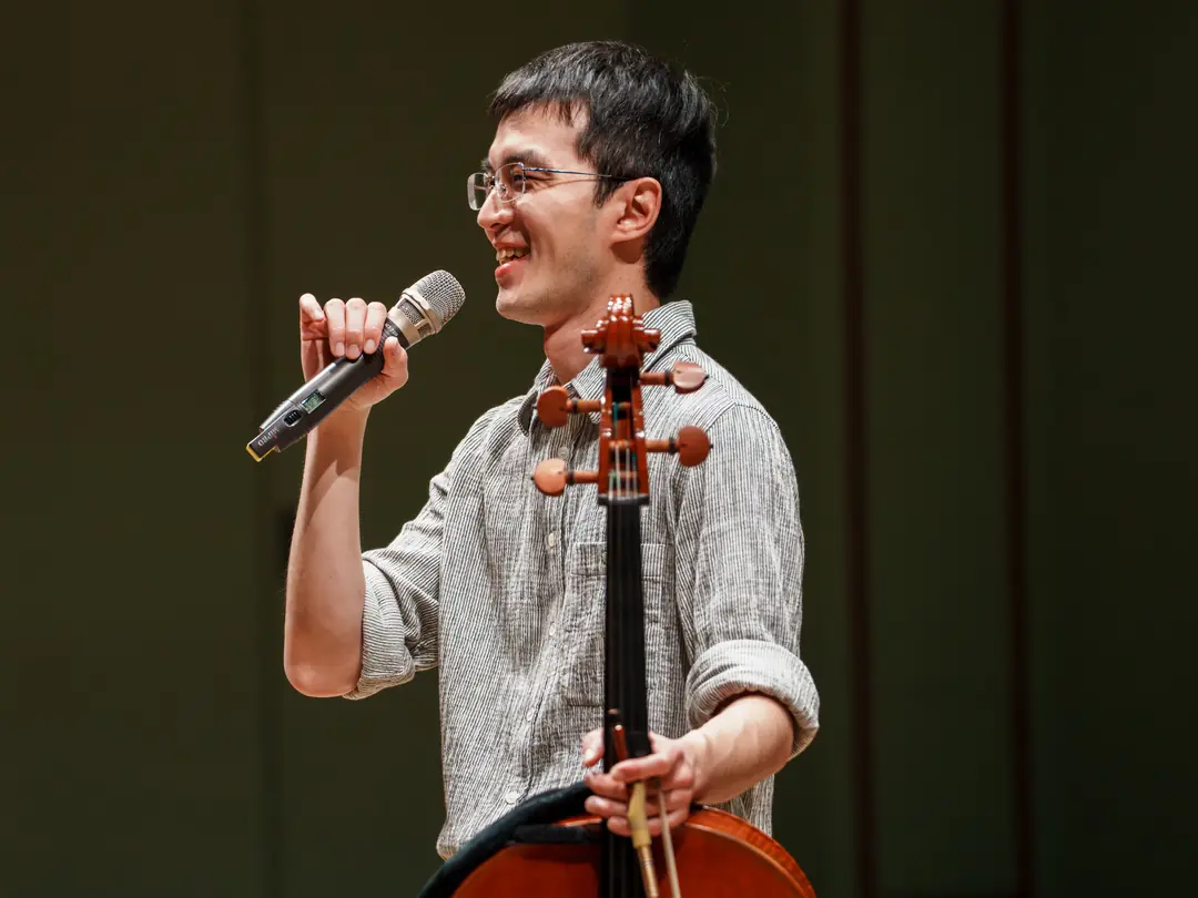 Dr. Su on stage holding a cello and speaking into a microphone